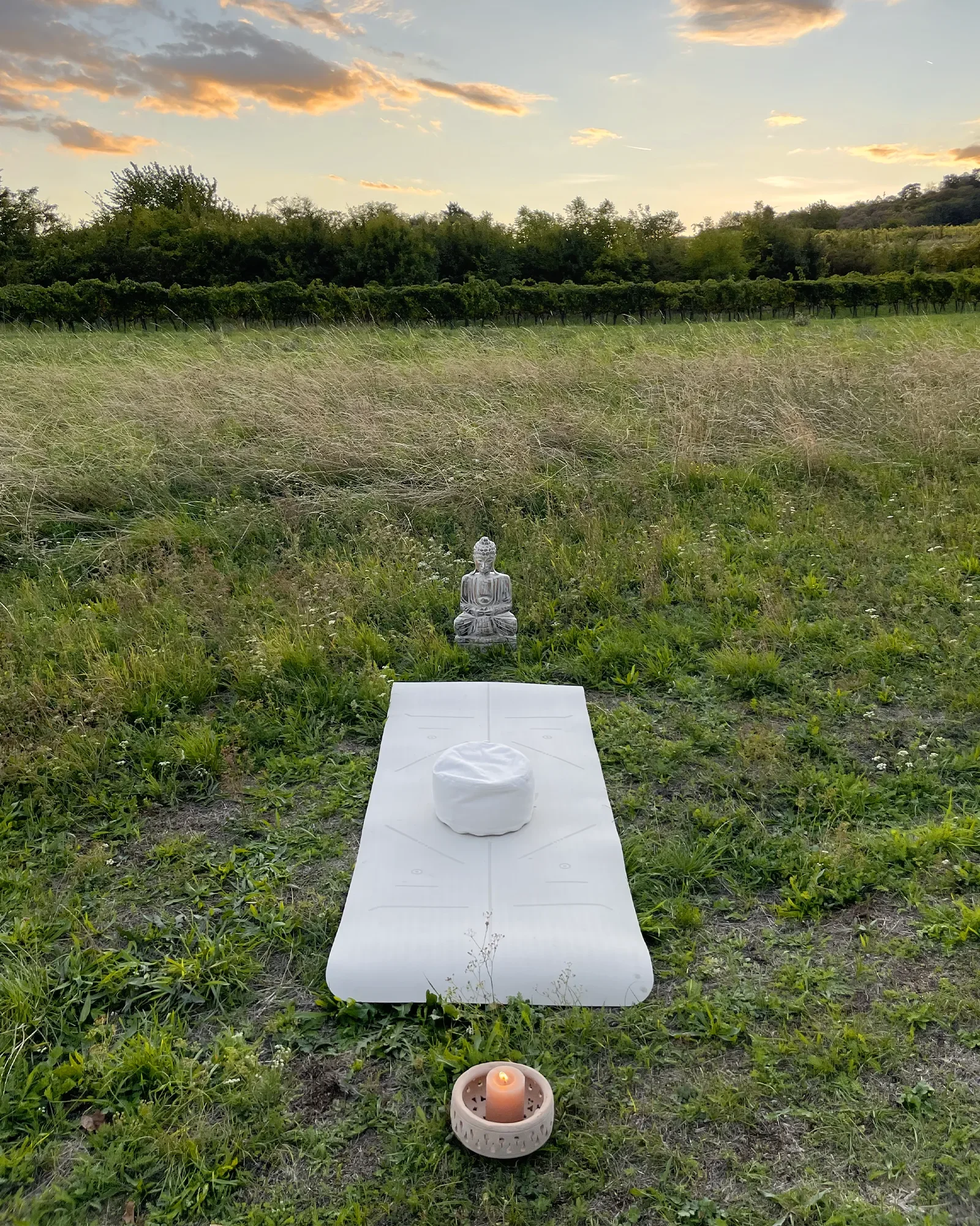 Yogamatte mit Sitzkissen, Buddha-Figur und Kerze auf einer Wiese im Abendlicht